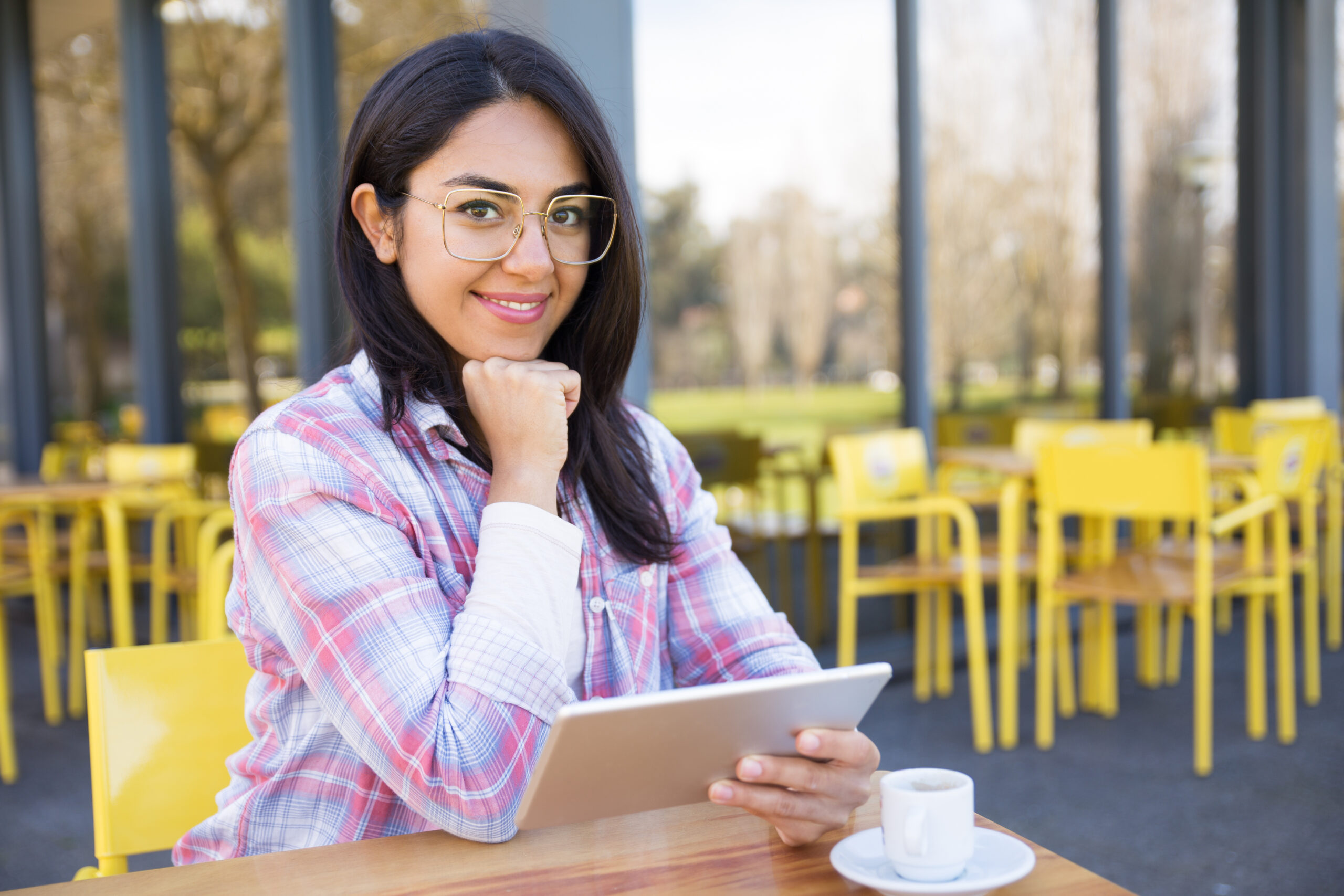 Smiling woman using tablet and drinking coffee in cafe. Pretty young lady wearing casual shirt and sitting with chairs and glass wall in background. Leisure and technology concept. Front view.